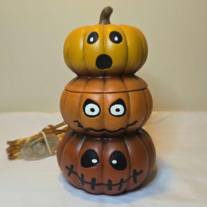 May include: A ceramic lamp shaped like a stack of three orange pumpkins with carved faces. The top pumpkin has a surprised expression, the middle has an angry face, and the bottom has a stitched mouth. Includes a brown electrical cord.