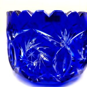 Op de afbeelding: Een diepblauwe, geslepen glazen kom met een geschulpte rand. De kom heeft een sterren- en geometrisch patroon, dat licht reflecteert. Het glas heeft een briljante glans en de kom staat tegen een witte achtergrond. Een decoratief stuk.
