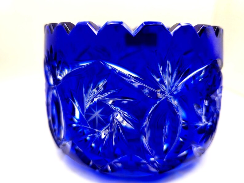 Op de afbeelding: Een diepblauwe, geslepen glazen kom met een geschulpte rand. De kom heeft een sterren- en geometrisch patroon, dat licht reflecteert. Het glas heeft een briljante glans en de kom staat tegen een witte achtergrond. Een decoratief stuk.
