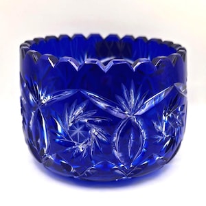 Op de afbeelding: Een diepblauwe, geslepen glazen kom met een geschulpte rand. De kom heeft ingewikkelde patronen van sterren en bloemen, waardoor een sprankelend effect ontstaat. Deze decoratieve kom is ideaal om te serveren of tentoon te stellen.