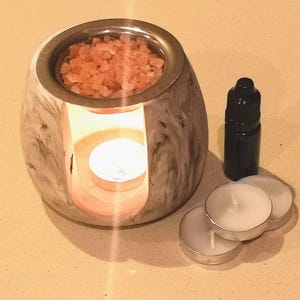 May include: A marble-effect essential oil diffuser with a metal rim, filled with pink salt crystals. A small black bottle and three tea light candles are next to the diffuser. A lit candle is inside.