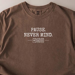 Puede incluir: Camiseta color espresso con el texto blanco "PAUSE. NEVER MIND.". La camiseta tiene cuello redondo y mangas cortas. El texto está centrado en el pecho, con una línea y símbolos debajo. Hecha de un material suave y cómodo.