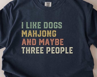May include: True navy blue t-shirt with the text "I LIKE DOGS MAHJONG AND MAYBE THREE PEOPLE" in a retro font. The shirt is a Comfort Colors brand. The text is in green, yellow, and red.