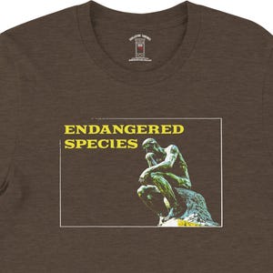 May include: Brown t-shirt featuring the text "ENDANGERED SPECIES" above an image of a statue. The statue is green and depicts a person in a thinking pose. The t-shirt is made of a soft material.