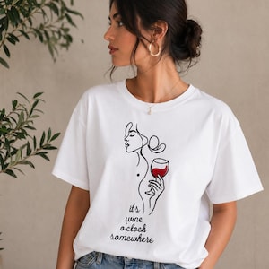 Könnte beinhalten: Weißes T-Shirt mit einer schwarzen Strichzeichnung einer Frau, die ein Weinglas mit Rotwein hält. Der Text "it's wine o'clock somewhere" befindet sich darunter. Die Person, die das T-Shirt trägt, hat dunkles Haar und goldene Creolen.