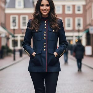 Op de afbeelding: Marineblauwe jas met gouden knopen en rode bies. De jas heeft een hoge kraag en vier zakken. De jas wordt gedragen met een donkere broek. Het ontwerp van de jas is militair geïnspireerd.