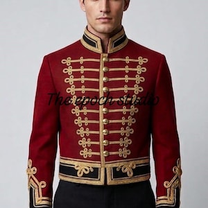 Pode incluir: Uma jaqueta de estilo militar em vermelho escuro com gola alta, botões dourados e detalhes intrincados de cordão trançado dourado na frente. A peça inclui acabamentos pretos nos punhos e na bainha, com o texto The epoch studio visível.