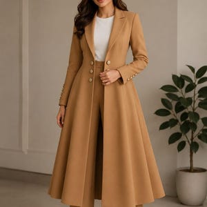 Peut inclure: Un tailleur-pantalon pour femme de couleur beige, composé d'un long manteau à jupe évasée et boutons dorés, assorti d'un pantalon large. Le manteau est ajusté et à manches longues. Un haut blanc est visible en dessous. Idéal pour les occasions formelles.