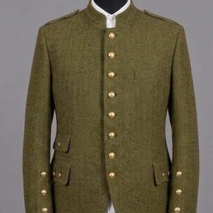 Puede incluir: Chaqueta de tweed verde oliva con diseño de espiga, cuello mandarín y botones dorados. La chaqueta tiene dos bolsillos delanteros y botones dorados en las mangas. La chaqueta se exhibe en un maniquí negro.