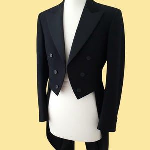 Op de afbeelding: Een formele zwarte smokingjas met een dubbele rij knopen en een revers. De jas heeft een klassiek staartjasontwerp, met een lange achterkant en een kortere voorkant. De jas wordt getoond op een witte mannequin tegen een gele achtergrond.