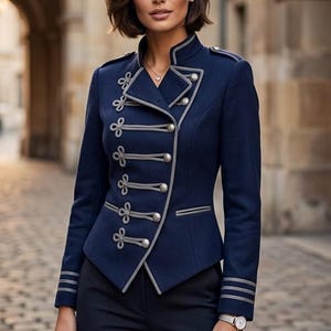May include: A navy blue military-style jacket featuring a high collar, silver-toned buttons, and decorative grey braided cord detailing. The jacket has long sleeves with three horizontal grey stripes at the cuffs and is paired with dark trousers.