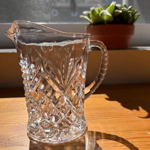 May include: A clear, vintage-style glass creamer with a diamond-cut pattern. The creamer has a handle and a spout for easy pouring. The glass is sitting on a wooden surface, with a shadow cast by the sunlight.