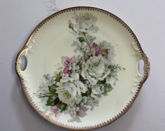 May include: A round, cream-colored serving platter with a floral design. The platter features a gold trim and handles. The floral arrangement includes white roses, pink flowers, and green leaves. A decorative piece for serving food.