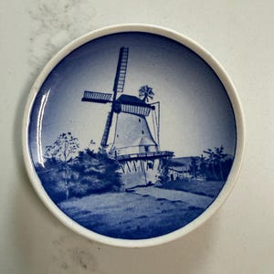 Puede incluir: Plato decorativo pequeño, redondo, azul y blanco, con una escena de molino de viento. El molino es blanco con detalles azules, sobre un cielo y paisaje azules. El plato tiene un borde blanco y una estética vintage clásica.