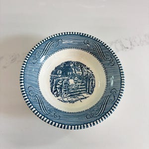 May include: A blue and white ceramic bowl with a detailed village scene in the center. The rim features a decorative pattern with a scalloped edge. The bowl is likely vintage and suitable for serving soup or cereal.