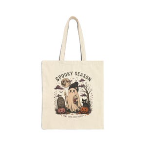 Peut inclure: Un sac fourre-tout en toile beige avec une illustration graphique d'un fantôme portant un bonnet noir et tenant une boisson. Le motif comprend des citrouilles, une pierre tombale, un chat noir, un arbre et le texte SPOOKY SEASON et STAY CUTE, STAY CREEPY.