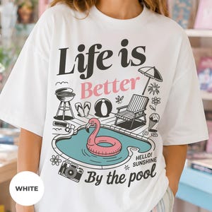 Op de afbeelding: Een wit t-shirt met een grafische print met de tekst Life is Better By the pool en Hello! Sunshine. Het ontwerp bevat een zwembad met een roze flamingo-zwemband, een barbecue, een strandstoel, een strandbal, slippers en een fotocamera.