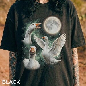 Pode incluir: T-shirt preta com um gráfico de três gansos brancos com bicos e patas laranja, contra um céu estrelado e uma lua cheia. A palavra "BLACK" está impressa na parte inferior da camisa.