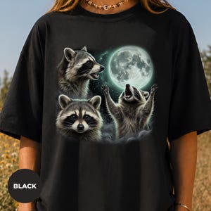 Può includere: Una t-shirt nera con una stampa grafica di tre procioni. Un procione guarda in avanti, uno è di profilo e uno tende le zampe verso una grande luna piena luminosa circondata da una nebbia soffusa ed eterea e un cielo stellato.