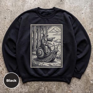 May include: A black crewneck sweatshirt featuring a rectangular graphic print of a wizard in a pointed hat riding a large snail through a forest. The illustration is rendered in a black and white woodcut style, showing the wizard pointing forward.