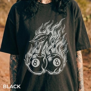 Peut inclure: T-shirt noir avec un motif de deux cerises avec le chiffre 8, entourées de flammes. Le texte "BLACK" est en bas de l'image. Le t-shirt a des manches courtes et un col rond.