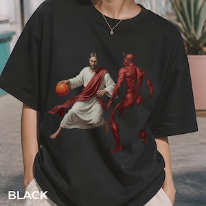 Könnte beinhalten: Schwarzes T-Shirt mit einer Grafik von Jesus, der Basketball gegen einen roten Teufel spielt. Jesus trägt ein weißes Gewand und einen roten Schärpen, während der Teufel rot ist. Das Wort "BLACK" ist unten aufgedruckt.