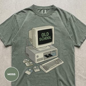 May include: A moss green short-sleeve t-shirt featuring a graphic of a vintage beige computer system. The monitor displays the text OLD SCHOOL in pixelated font. A keyboard and several floppy disks are illustrated below the computer tower.
