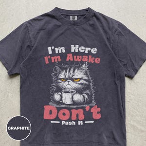 Könnte beinhalten: Ein anthrazitfarbenes Kurzarm-T-Shirt mit einer Grafik einer mürrischen Katze, die eine Kaffeetasse hält. Der Text lautet I'm Here I'm Awake Don't Push It. Die Katze hat gelbe Augen und dunkles Fell. Das T-Shirt hat einen Rundhalsausschnitt und eine verwaschene Optik.