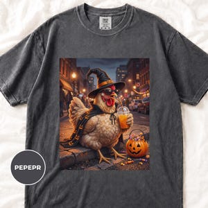 May include: A charcoal gray t-shirt featuring a graphic of a chicken wearing a black witch hat, sunglasses, and a cape with pumpkin patterns. The chicken holds an orange drink in a plastic cup next to a jack-o'-lantern bucket filled with candy.