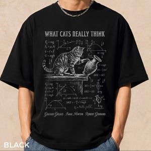 Könnte beinhalten: Schwarzes T-Shirt mit dem Text "WHAT CATS REALLY THINK" und einer Grafik einer Katze, Gleichungen und wissenschaftlichen Diagrammen. Das Shirt zeigt die Namen Galileo Galilei, Isaac Newton und Robert Goddard.