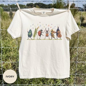 Può includere: Una t-shirt color avorio a maniche corte con una stampa grafica di sei insetti che suonano strumenti musicali. L'illustrazione include uno scarabeo, una cavalletta, una coccinella, un cervo volante, un'ape e un altro scarabeo con note musicali.