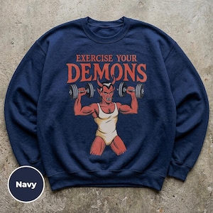 May include: A navy blue crewneck sweatshirt featuring a graphic of a red devil with horns wearing a white tank top and lifting grey dumbbells. The text above the figure reads EXERCISE YOUR DEMONS in a bold, orange-red font. The fabric is solid.
