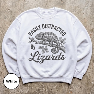 Puede incluir: Una sudadera blanca de cuello redondo con un gráfico gris oscuro de un camaleón posado en una rama. El texto "EASILY DISTRACTED By Lizards" está impreso en una combinación de fuentes de bloque y cursiva alrededor de la ilustración.