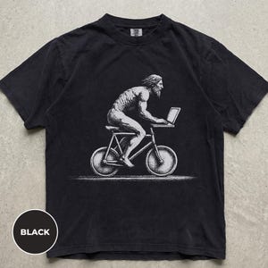 Può includere: Una t-shirt nera a maniche corte con una stampa grafica bianca raffigurante una figura muscolosa e barbuta in bicicletta mentre usa un computer portatile. Il design è realizzato in uno stile illustrativo puntinato, centrato sul petto del capo.