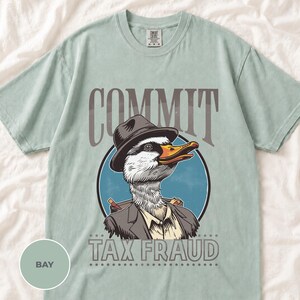 May include: A light sage green t-shirt featuring a graphic of a goose wearing a fedora and a suit jacket. The text above the goose reads COMMIT and the text below reads TAX FRAUD. The design is set within a circular blue background.