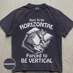 May include: A charcoal gray short-sleeve t-shirt featuring a white graphic of a tabby cat lying on a pillow. The text above the cat reads Born To Be HORIZONTAL and the text below reads FORCED to BE VERTICAL. A circular label reads GRAPHITE.