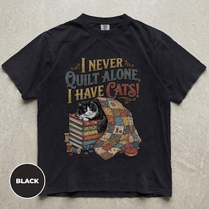 Puede incluir: Una camiseta negra de manga corta con un gráfico de un gato blanco y negro sobre una pila de libros bajo una colcha de retazos colorida. El texto dice, I NEVER QUILT ALONE, I HAVE CATS! Se ven carretes de hilo y un alfiletero.