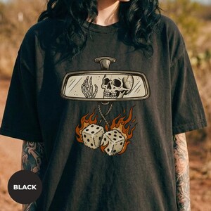 May include: A black t-shirt featuring a graphic print of a car rearview mirror containing a skeleton hand and skull. Hanging from the mirror are two dice surrounded by orange and yellow flames, rendered in a detailed, illustrative style.