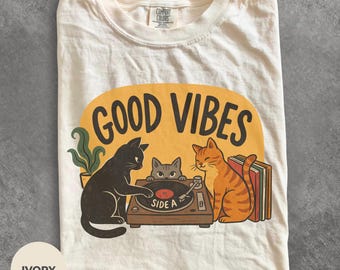 May include: An ivory t-shirt featuring a graphic of three cats around a record player. The text GOOD VIBES is printed above the cats. One black cat touches the record, a gray cat peeks over the player, and an orange tabby cat sits nearby.