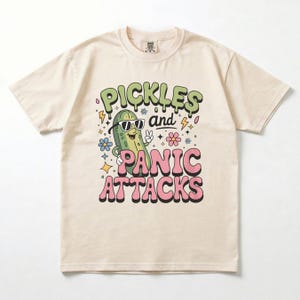 Könnte beinhalten: Cremefarbenes T-Shirt mit einer Cartoon-Gurke, die eine Sonnenbrille trägt und ein Friedenszeichen zeigt. Das Shirt trägt die Worte "PICKLES and PANIC ATTACKS" in grünen und rosa Buchstaben, mit dekorativen Blumen und Blitzen.