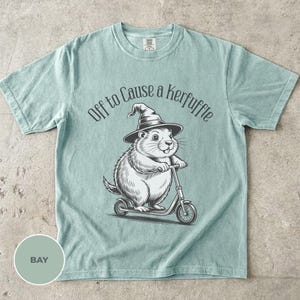 May include: A light green short-sleeve t-shirt featuring a black and white graphic of a groundhog wearing a witch hat and riding a kick scooter. Above the illustration, the text reads "Off to Cause a Kerfuffle" in a curved, dark font.