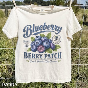 May include: An ivory t-shirt featuring a graphic of blueberries with green leaves. The text reads Blueberry Berry Patch, Maine 1986, Fresh Picked, and Small Berries Big Summer. The design uses blue and green tones on a light-colored fabric background.