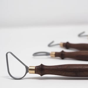 Puede incluir: Cuatro herramientas de bucle de cerámica con mangos de madera marrón oscuro y férulas de latón. Las herramientas tienen un bucle de metal gris en el extremo. Las herramientas están dispuestas sobre una superficie blanca.