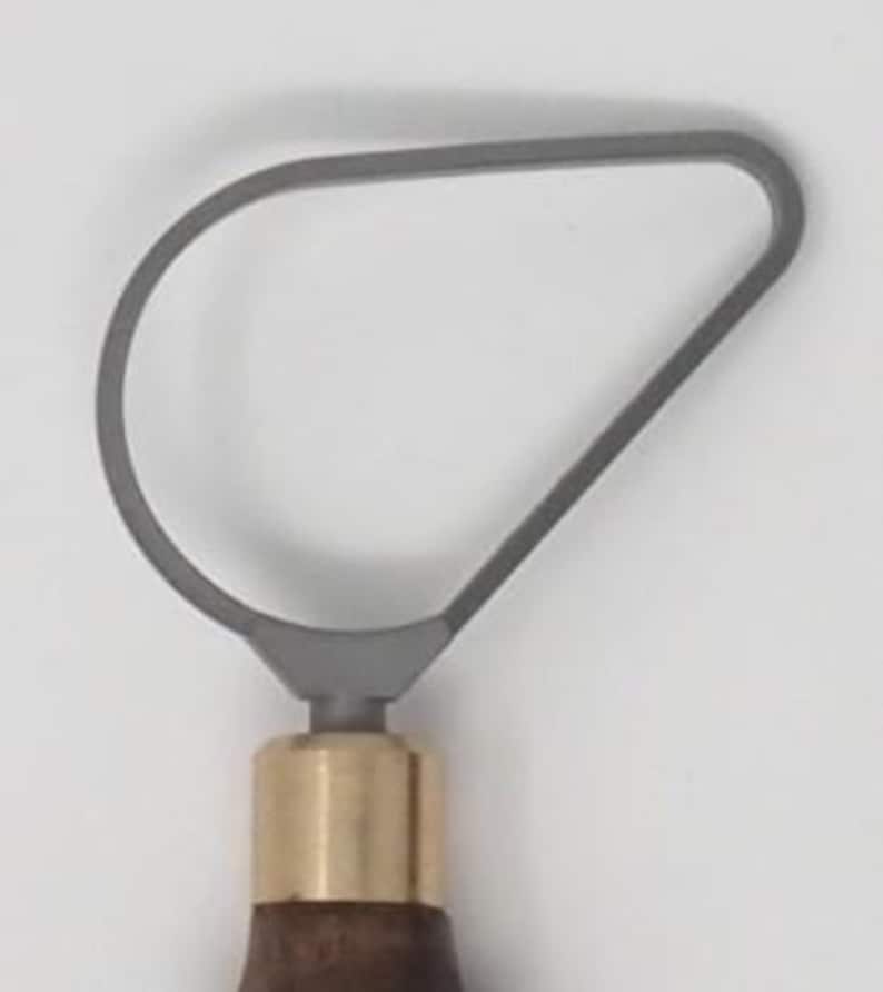 May include: A gray loop tool for pottery or sculpting, featuring a curved metal loop attached to a wooden handle with a brass ferrule. The tool is designed for shaping and removing clay.