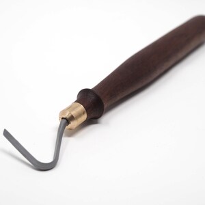 May include: A pottery tool with a dark brown wooden handle, a brass ferrule, and a gray, curved metal loop. The tool is designed for shaping and detailing clay. The handle is smooth and ergonomic.