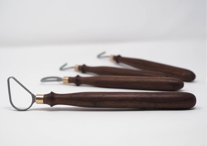 May include: Four pottery loop tools with dark brown wooden handles and brass ferrules. The loop tools have a gray metal loop at the end. The tools are arranged on a white surface.