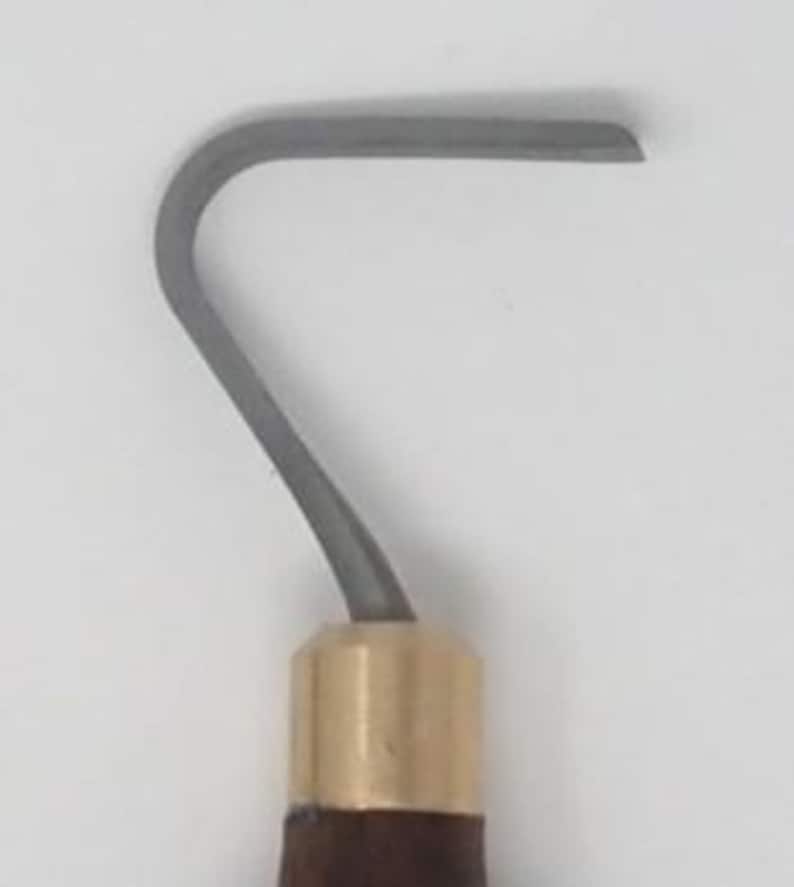 May include: A close-up of a gray, curved metal blade with a brass ferrule and a dark brown wooden handle. The blade is approximately 1 inch wide and has a rounded tip. The tool is likely used for sculpting or carving.