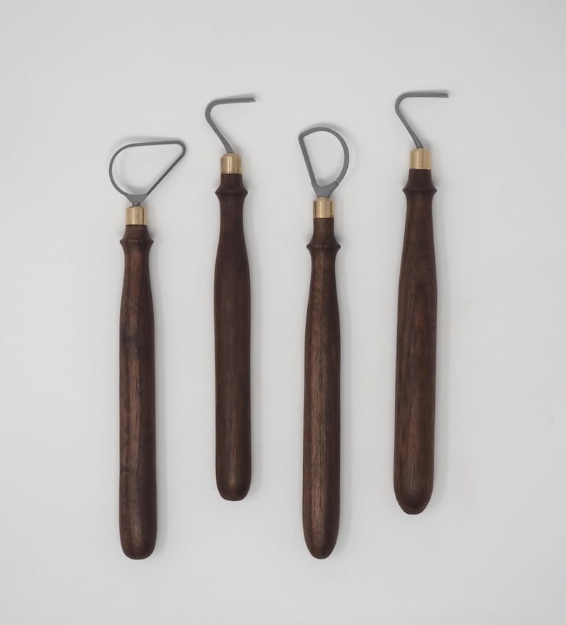 May include: Four pottery tools with dark brown wooden handles and brass-colored ferrules. The metal ends are gray and have different shapes, including a loop and a hook. The tools are arranged against a white background.