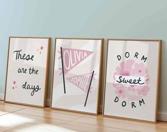 Può includere: Tre stampe artistiche incorniciate con testo e disegni. Una stampa dice "These are the days" con accenti rosa. Un'altra presenta gagliardetti con nomi. La terza dice "Dorm Sweet Dorm" con elementi floreali.