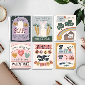 May include: A collection of six greeting cards with various illustrations and text. The cards feature designs such as a ghost with pumpkins, a rabbit with carrots, a rainbow with a house, and phrases like "Finals Suck But You Are Awesome."